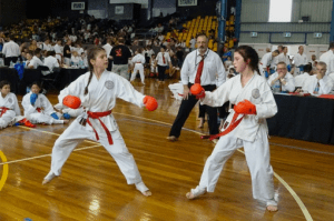 two young girls wearing a gi performing karate move wearing gloves in front of a crowd of karate students