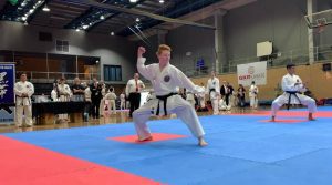 a young boy performs a karate stance, he wears a black belt, and there are observers around him