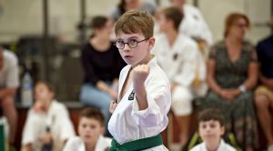 small boy wearing glasses, a gkr karate gi, and wearing a green belt performs a karate move