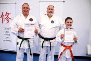 one older man stands in the middle of another older man holding a certificate, and a younger man holding a certificate wearing there karate gi
