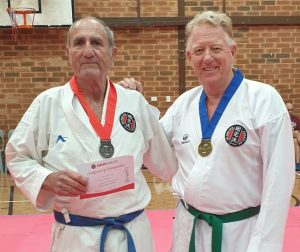 two older gentleman, one with a blue belt and one with a green, wear different medals and hold up a certificate of appreciation