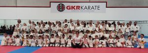 a large group of students wearing a karate gi in front of the gkr karate logo in a hall