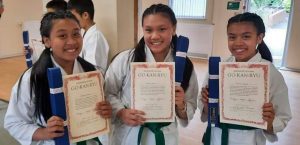 three young girls stand next to each other wearing a gi holding there brand new blue belts and a certificate of appreciation