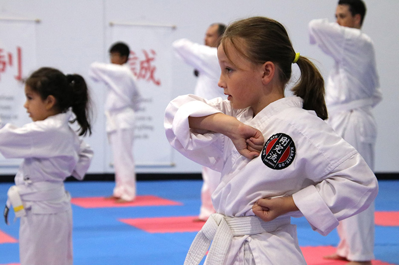 Young girl practicing karate in a dojo. She is wearing a white gi with a GKR Karate badge and a white belt. Other people can be seen training in the background.