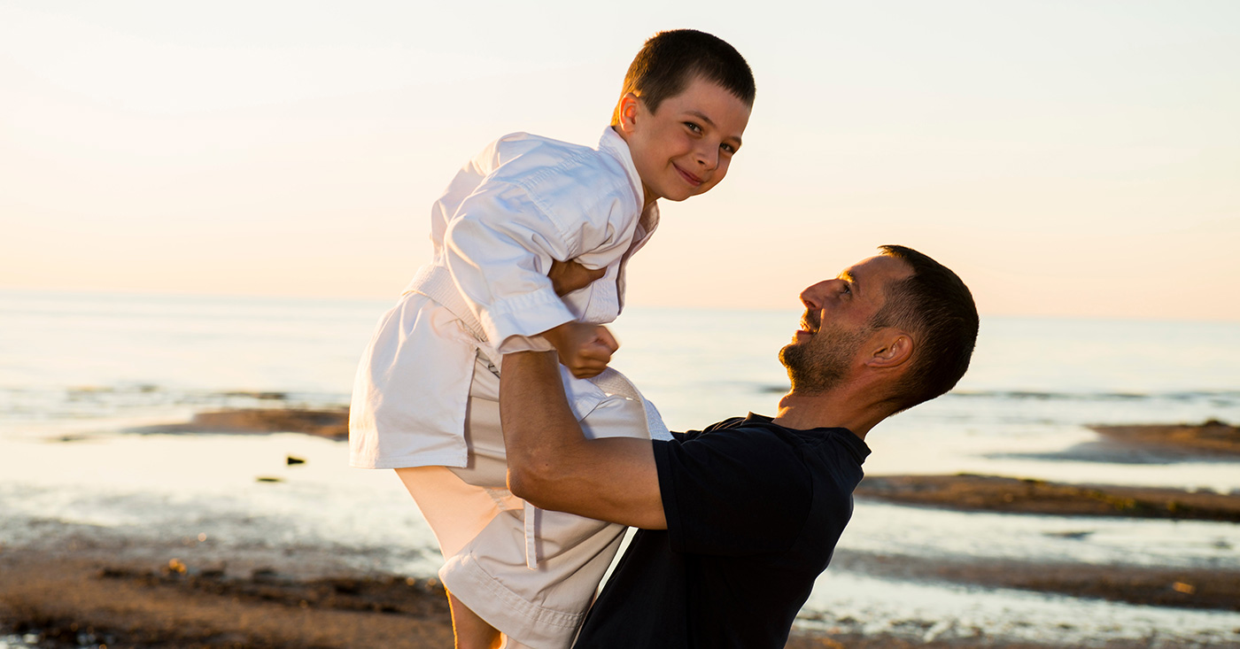 Father holding his son up in the air at the beach. The son is wearing a gi.