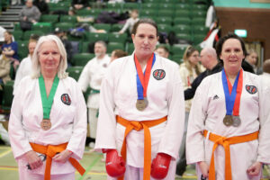 three women stand together, they are each wearing a gkr karate gi, and have medals around there neck