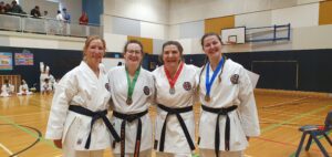 4 young women stand together with arms behind each others backs wearing karate gi at a basketball court