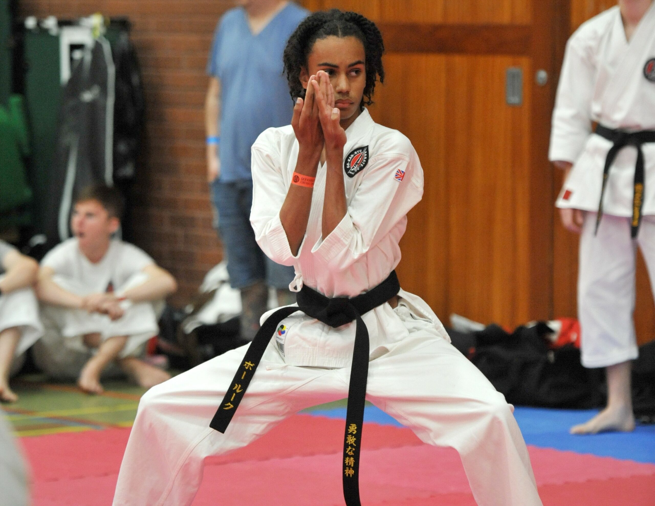 A focused young karateka with long hair is captured in a deep stance, performing a hand technique with intense concentration. Wearing a white gi and a black belt, the practitioner's gaze is directed forward, exuding calm and discipline. In the background, observers and fellow competitors are seated around the edge of the red and blue matted area of the dojo